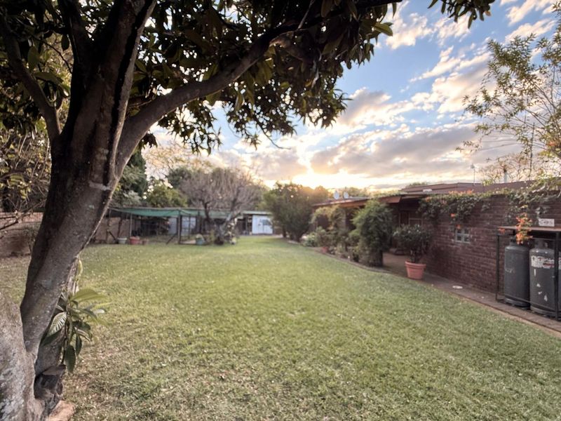 A spacious backyard featuring a well-maintained lawn, mature trees, and a brick boundary wall with an attached covered patio area.