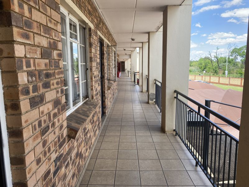 A long, covered outdoor balcony featuring tiled flooring, brick exterior walls, and metal balustrades overlooking the complex grounds.