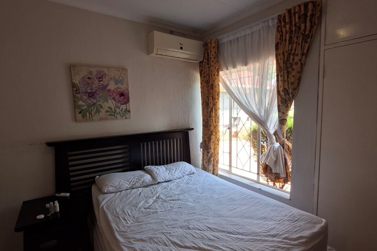 A functional bedroom featuring tiled flooring, a built-in cupboard, and a wall-mounted air conditioning unit, with natural light provided by a window with security bars.