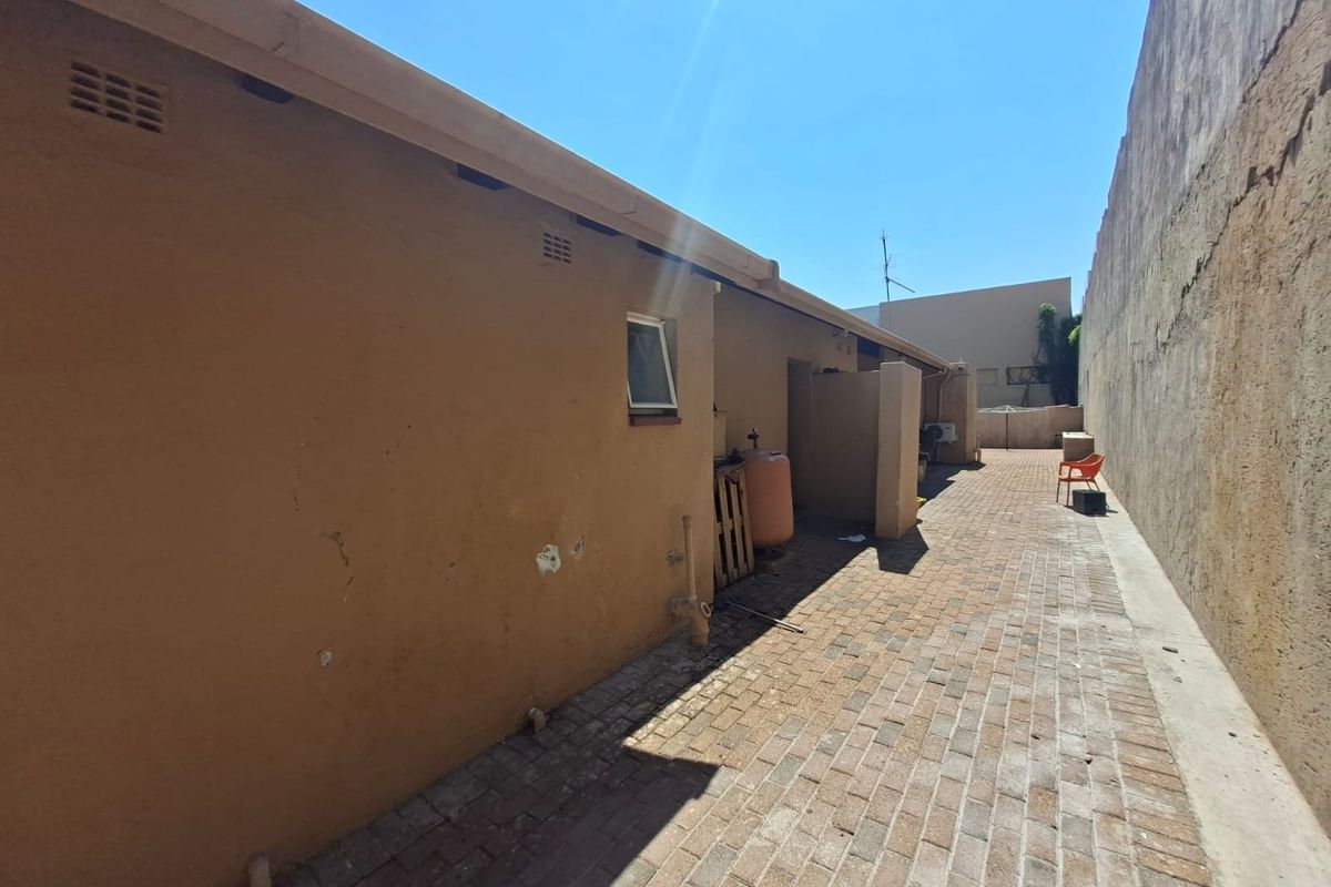 A narrow rear exterior side passage featuring brick paving, guttering, and a high boundary wall, showing signs of weathered exterior plaster.