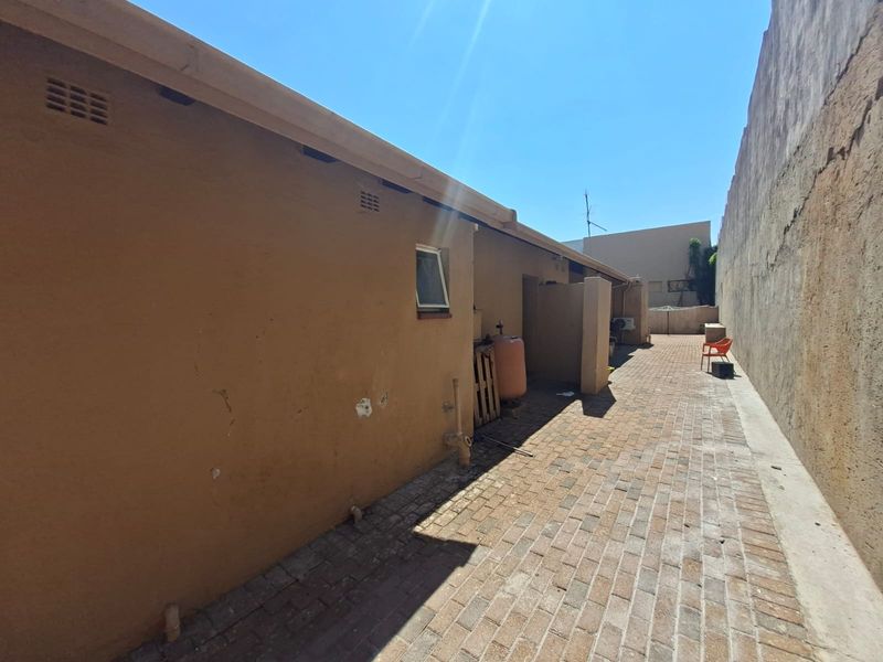 A narrow rear exterior side passage featuring brick paving, guttering, and a high boundary wall, showing signs of weathered exterior plaster.