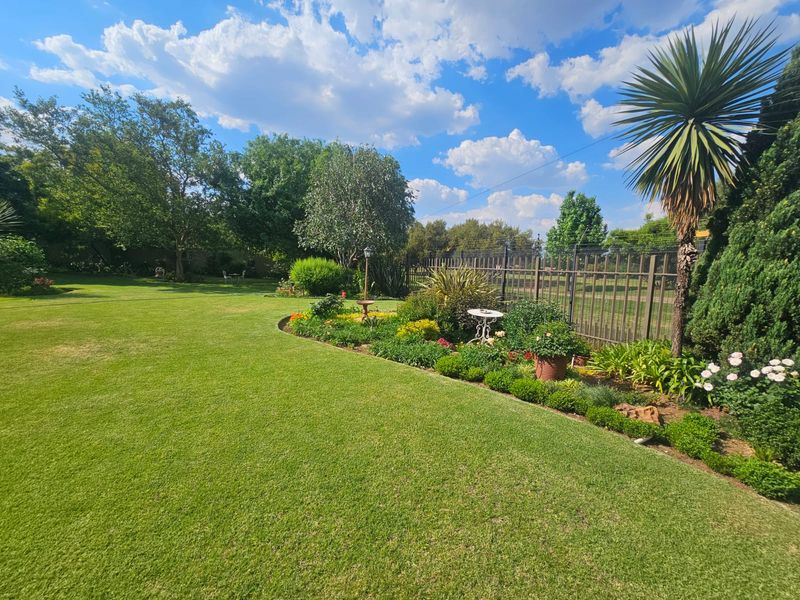 A well-maintained, spacious garden featuring a manicured lawn, established garden beds, and secure metal perimeter fencing under a clear blue sky.