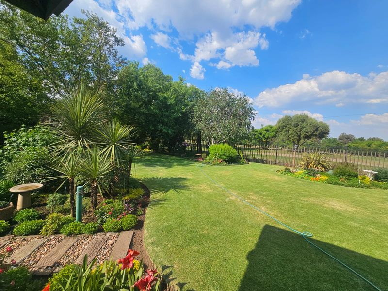 A well-maintained, landscaped garden featuring a lush green lawn, mature trees, and a stone-accented pathway enclosed by metal perimeter fencing.