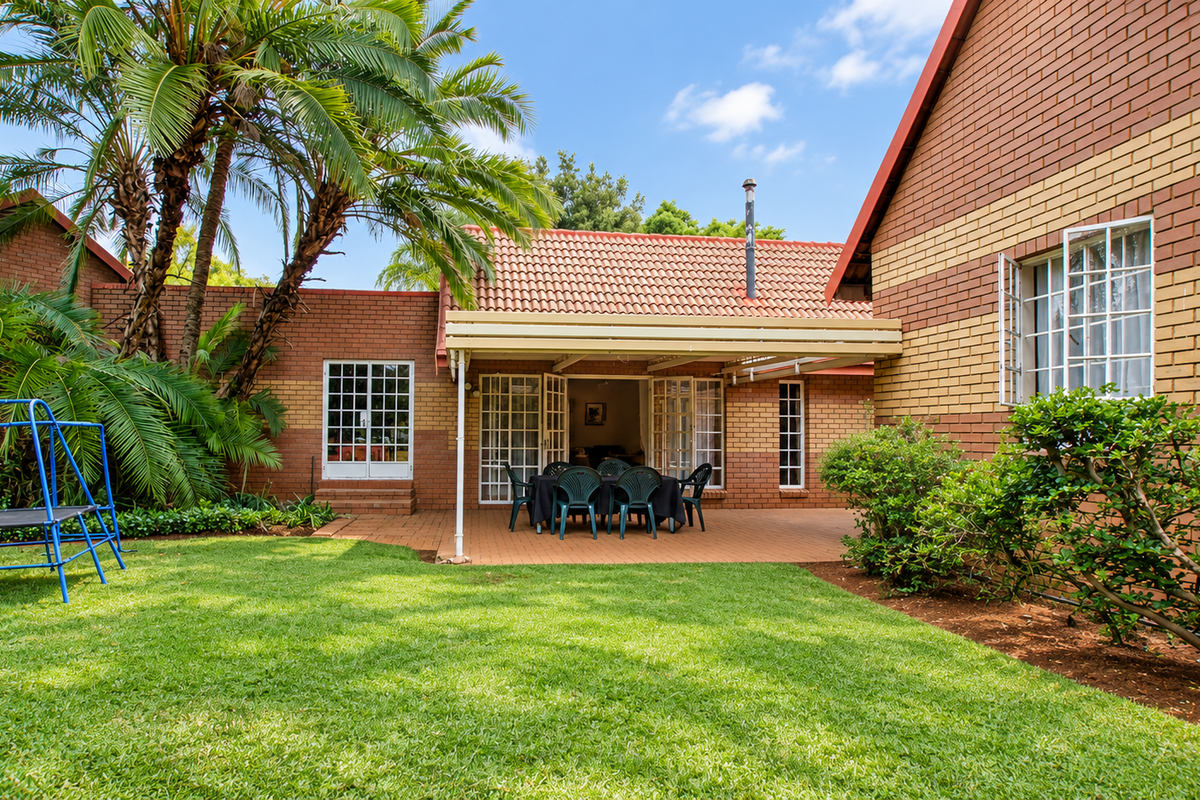 A well-maintained rear exterior view of the townhouse featuring a paved covered patio, brick facade, and a lush private garden.