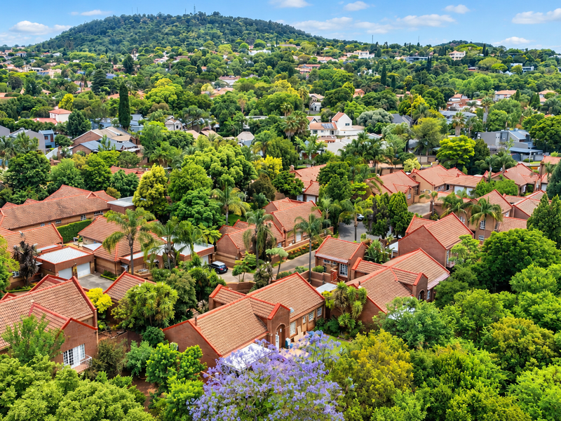 An elevated aerial view of a residential townhouse complex featuring brick buildings with tiled roofs, surrounded by lush green trees and established landscaping.
