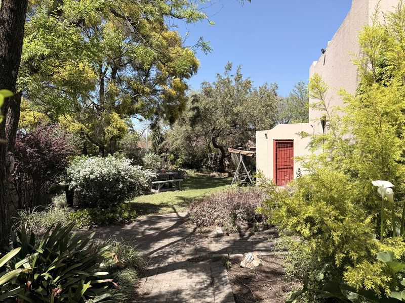 A lush, established garden featuring mature trees, landscaped shrubbery, and a paved pathway leading to a side entrance with a security gate.