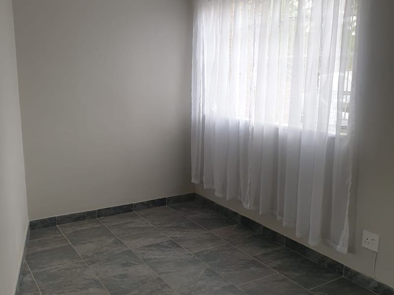 A clean, empty bedroom featuring neutral wall paint, grey tiled flooring, and a window fitted with a curtain rod.