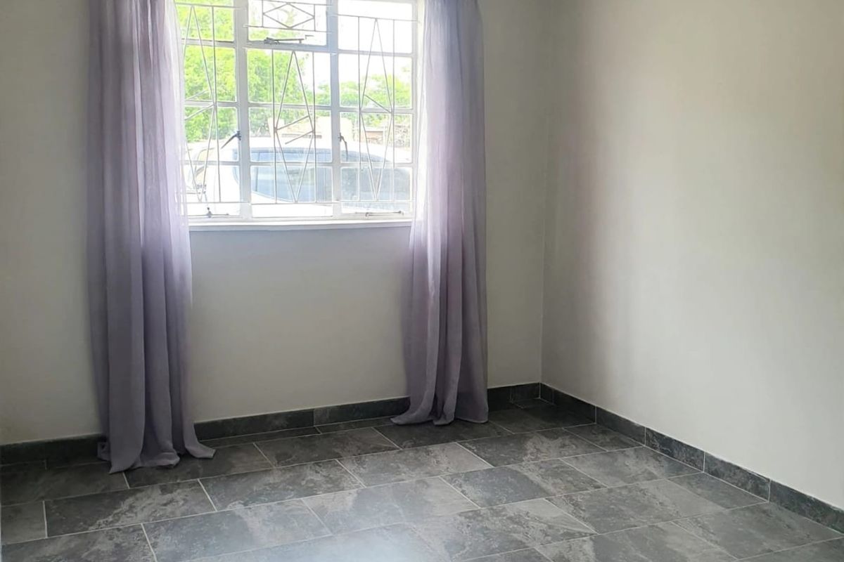 A bright, empty bedroom featuring neutral walls, modern grey tiled flooring, and a window fitted with security bars.