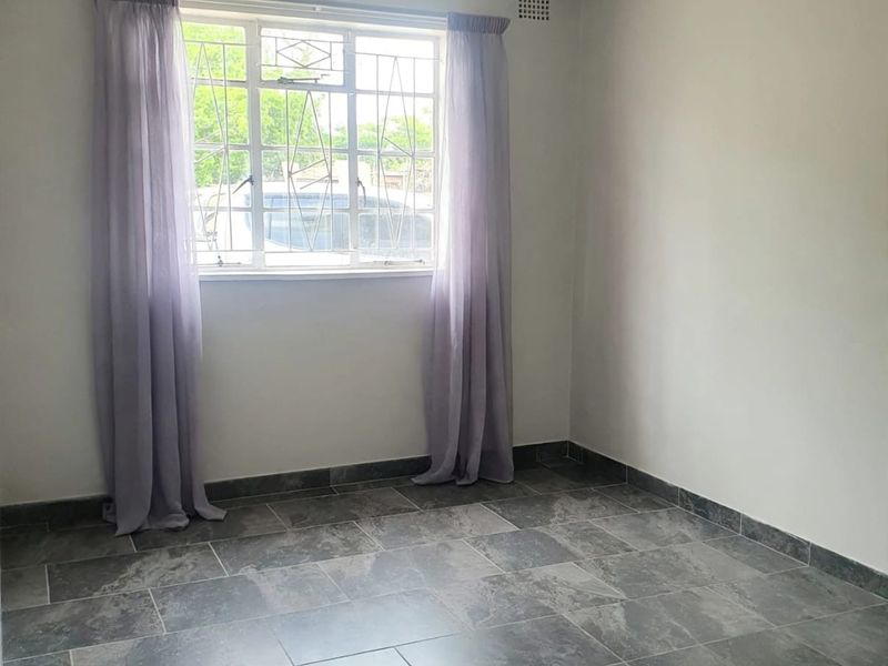 A bright, empty bedroom featuring neutral walls, modern grey tiled flooring, and a window fitted with security bars.