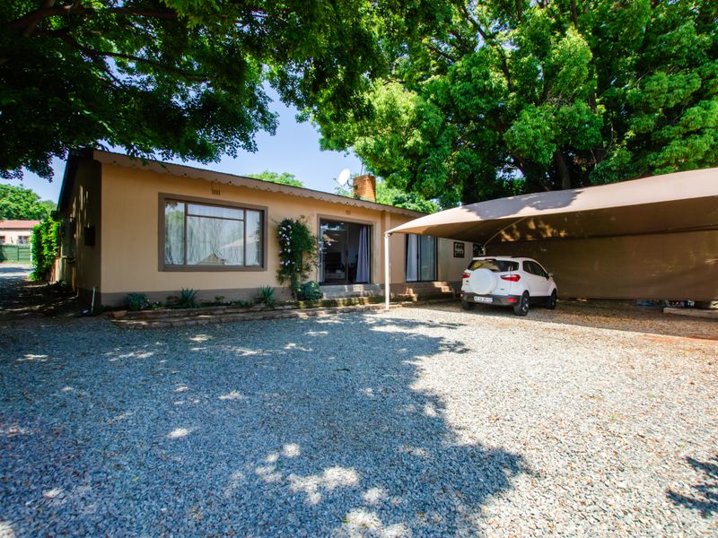 Front exterior view of a single-story house featuring a gravel driveway, a covered carport, and mature trees providing shade.