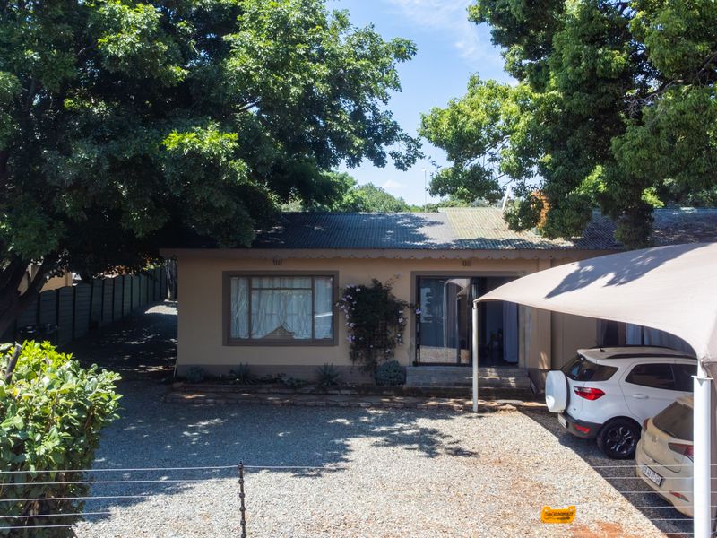 Front elevation of a single-story house featuring a gravel driveway, shade cloth carport, and mature trees providing ample shade.