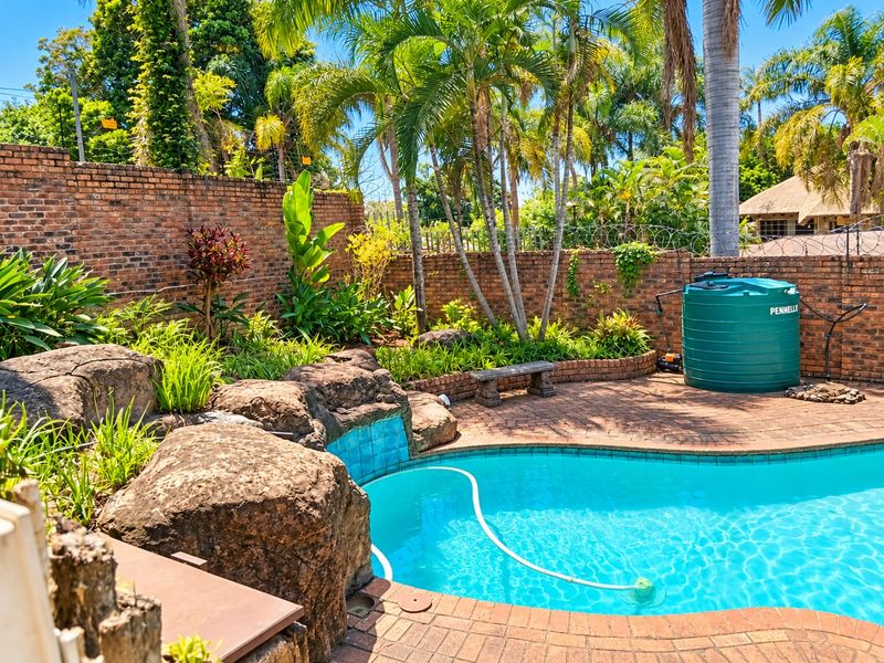 A private outdoor pool area featuring a paved deck, natural rock water feature, and a brick boundary wall with established garden landscaping.