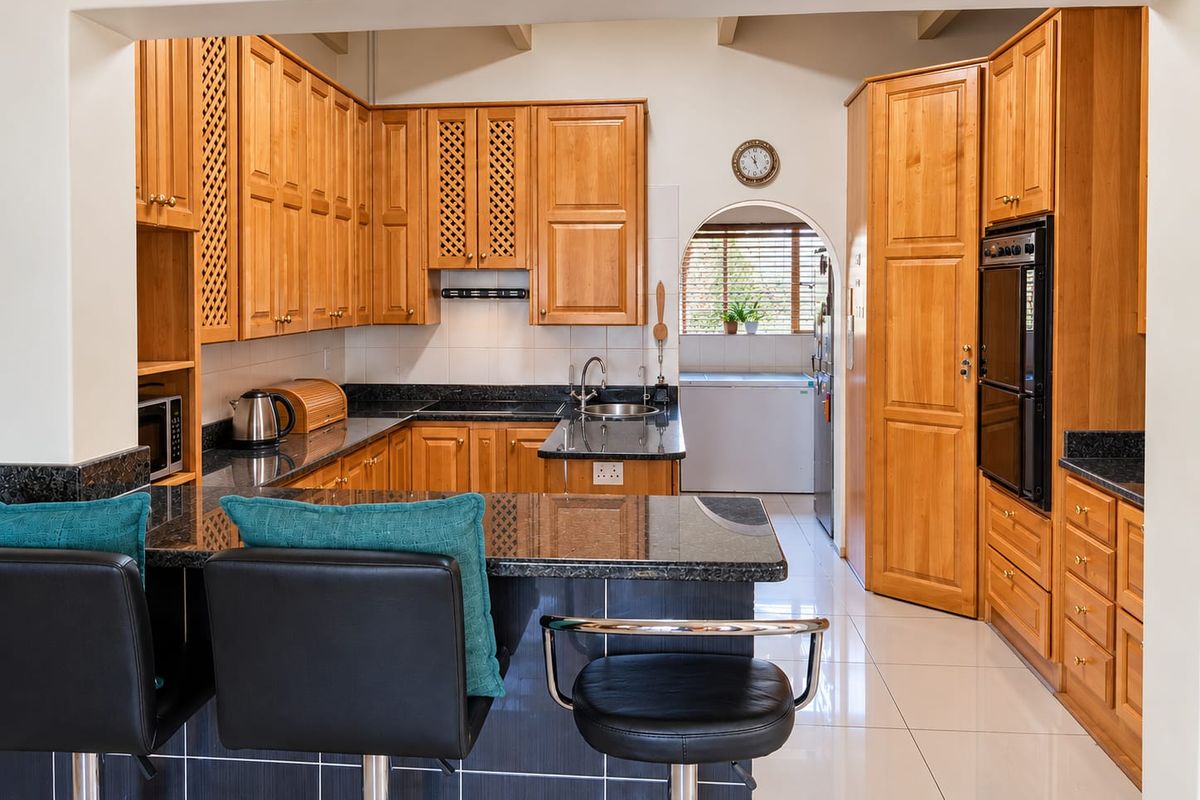 A well-maintained kitchen featuring warm wooden cabinetry, granite countertops, a breakfast bar, and integrated appliances, with a tiled floor and an adjacent scullery area.