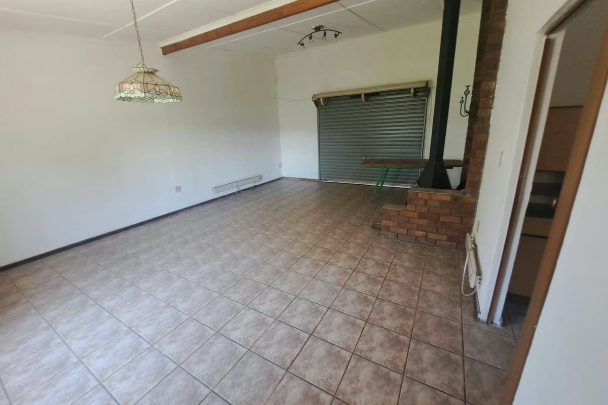 Spacious living area featuring tiled flooring, an exposed timber ceiling beam, and a brick-surround freestanding fireplace, with a roller shutter door leading outdoors.