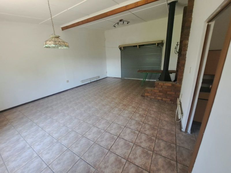 Spacious living area featuring tiled flooring, an exposed timber ceiling beam, and a brick-surround freestanding fireplace, with a roller shutter door leading outdoors.