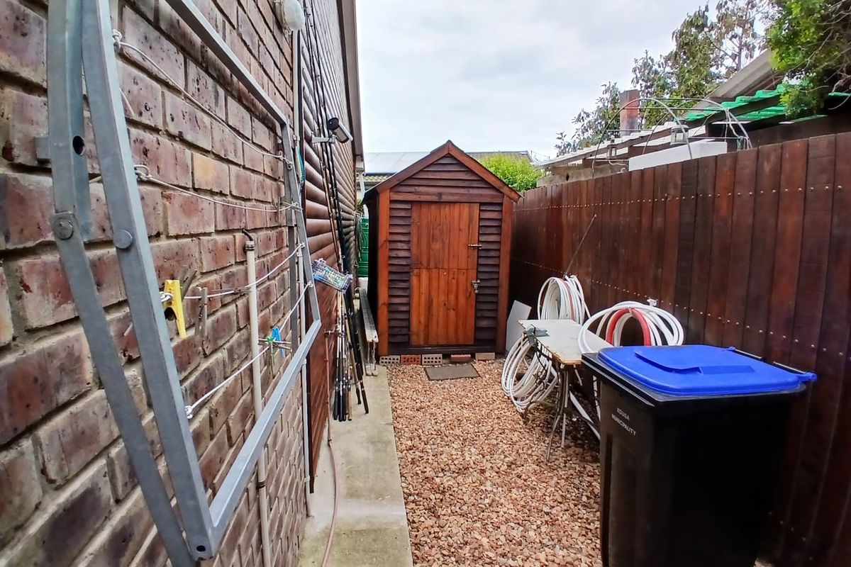 A side yard area featuring a brick wall, a timber storage shed, and a gravel pathway enclosed by a timber fence.