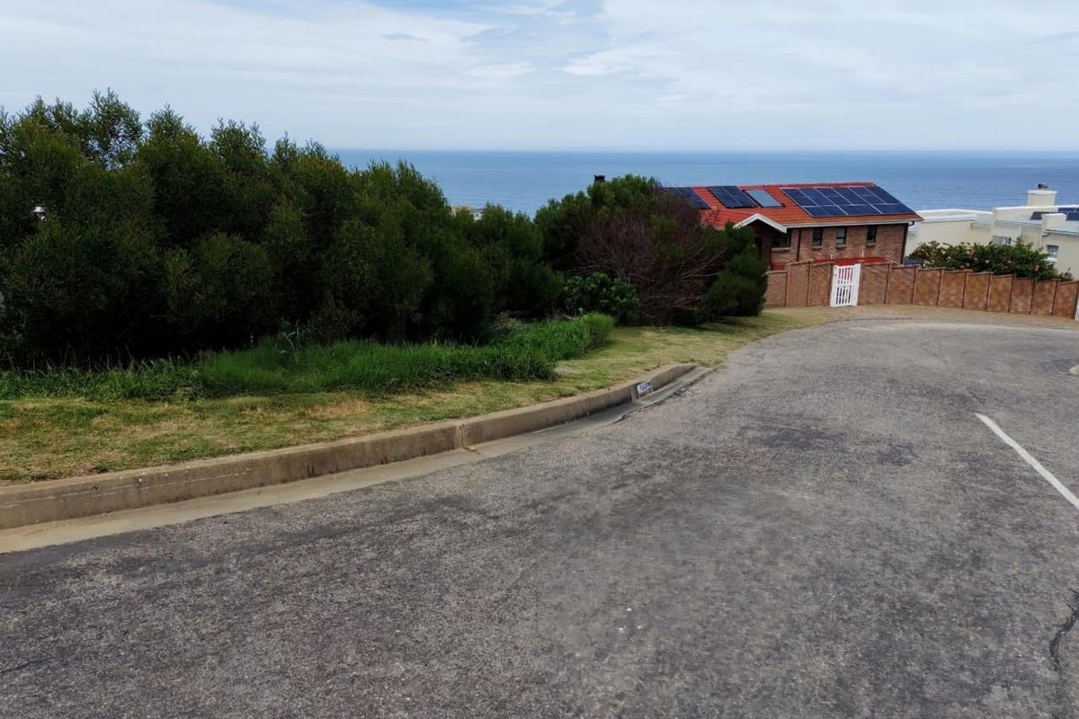 A street-level view of the residential plot featuring a paved road, concrete curbing, and a brick boundary wall with an ocean view in the background.