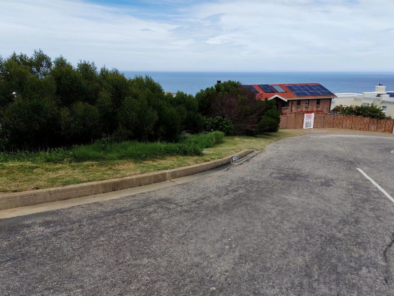 A street-level view of the residential plot featuring a paved road, concrete curbing, and a brick boundary wall with an ocean view in the background.