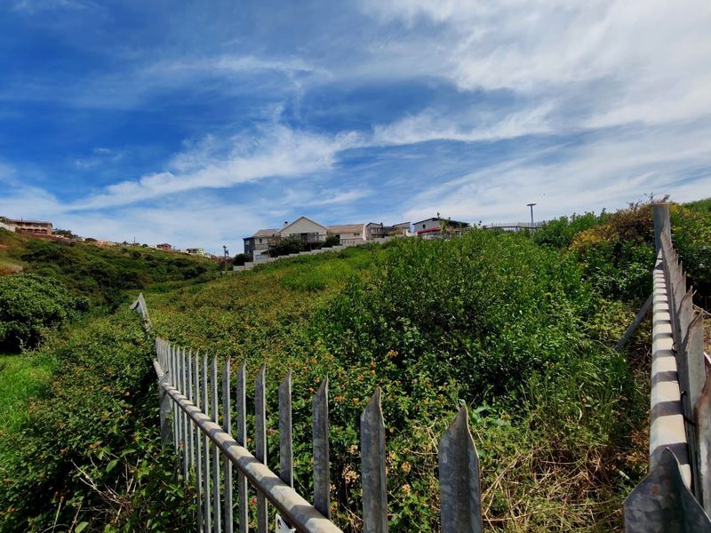 A view of a sloped, overgrown vacant residential plot featuring metal palisade fencing, with established residential properties visible in the background.