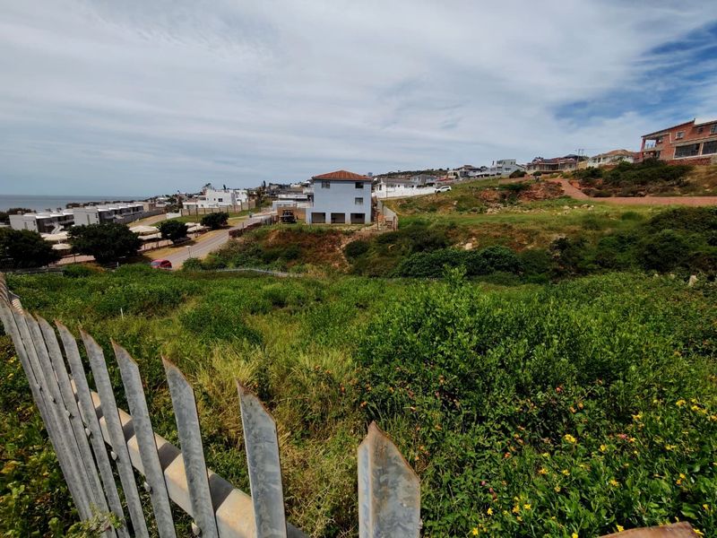 A view of a vacant residential plot featuring sloped terrain and metal palisade fencing, situated in a coastal neighborhood.