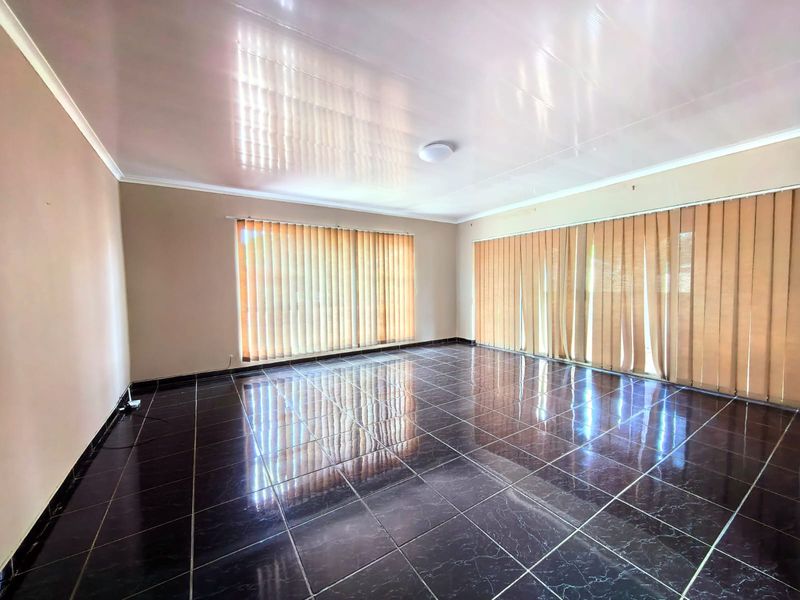 A spacious, empty living area featuring dark tiled flooring, neutral walls, and large windows fitted with vertical blinds.