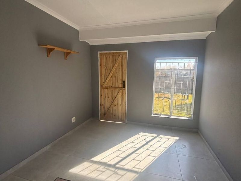 A vacant bedroom featuring tiled flooring, a wooden door, and a window fitted with security bars, offering a neutral canvas for personalization.