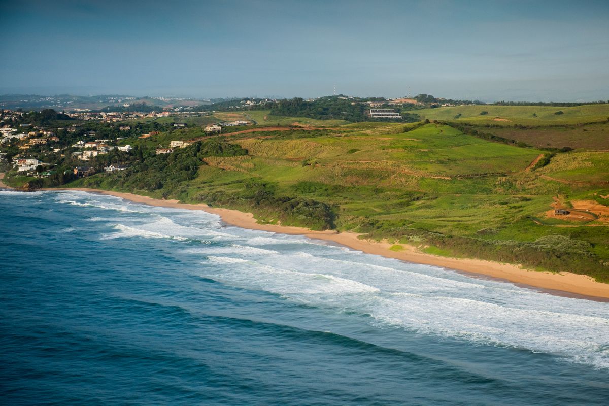 An aerial view of the coastline showing the ocean, sandy beach, and surrounding green hills.