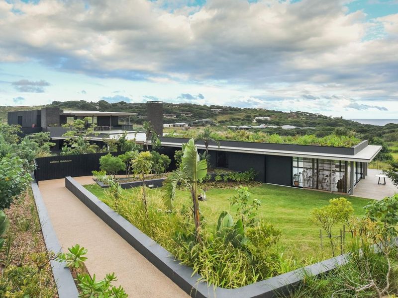 A modern architectural clubhouse featuring a green roof, landscaped gardens, and expansive glass facades overlooking a scenic coastal landscape.