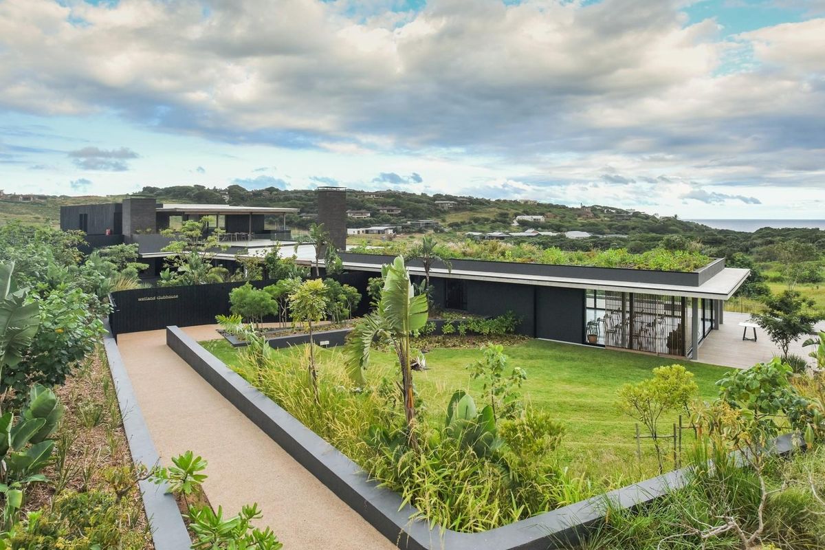 A modern architectural clubhouse featuring a green roof, landscaped gardens, and expansive glass facades overlooking a scenic coastal landscape.