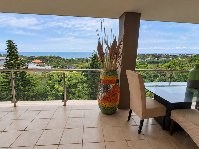 A spacious covered balcony featuring tiled flooring and a modern glass balustrade, offering expansive elevated views of the surrounding landscape and ocean.