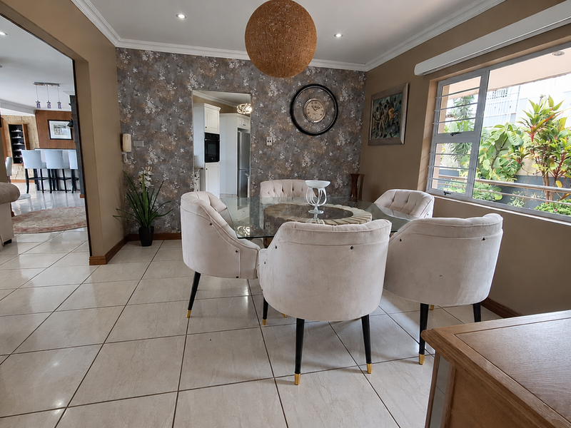 A bright dining room featuring tiled flooring, recessed lighting, and a feature wallpaper wall, with a view into the adjacent kitchen area.