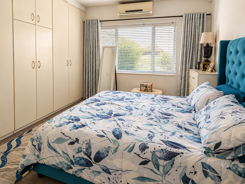 A well-maintained bedroom featuring extensive built-in cabinetry, a wall-mounted air conditioning unit, and large windows with venetian blinds and curtains.