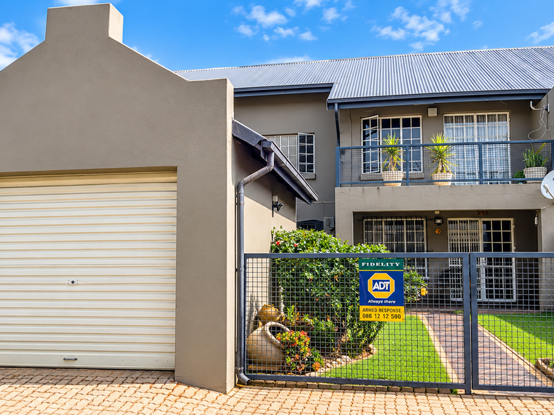Front elevation of a townhouse featuring a roller garage door, paved driveway, secure gated entrance, and an upper-level balcony with security bars.