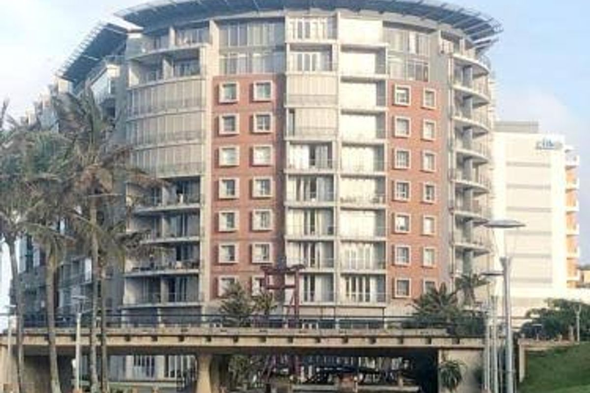 A view of a modern multi-story apartment building featuring a brick facade and balconies, situated along a canal with a pedestrian bridge.