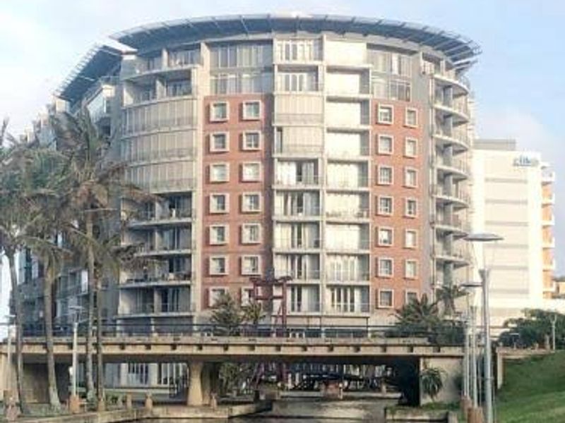 A view of a modern multi-story apartment building featuring a brick facade and balconies, situated along a canal with a pedestrian bridge.