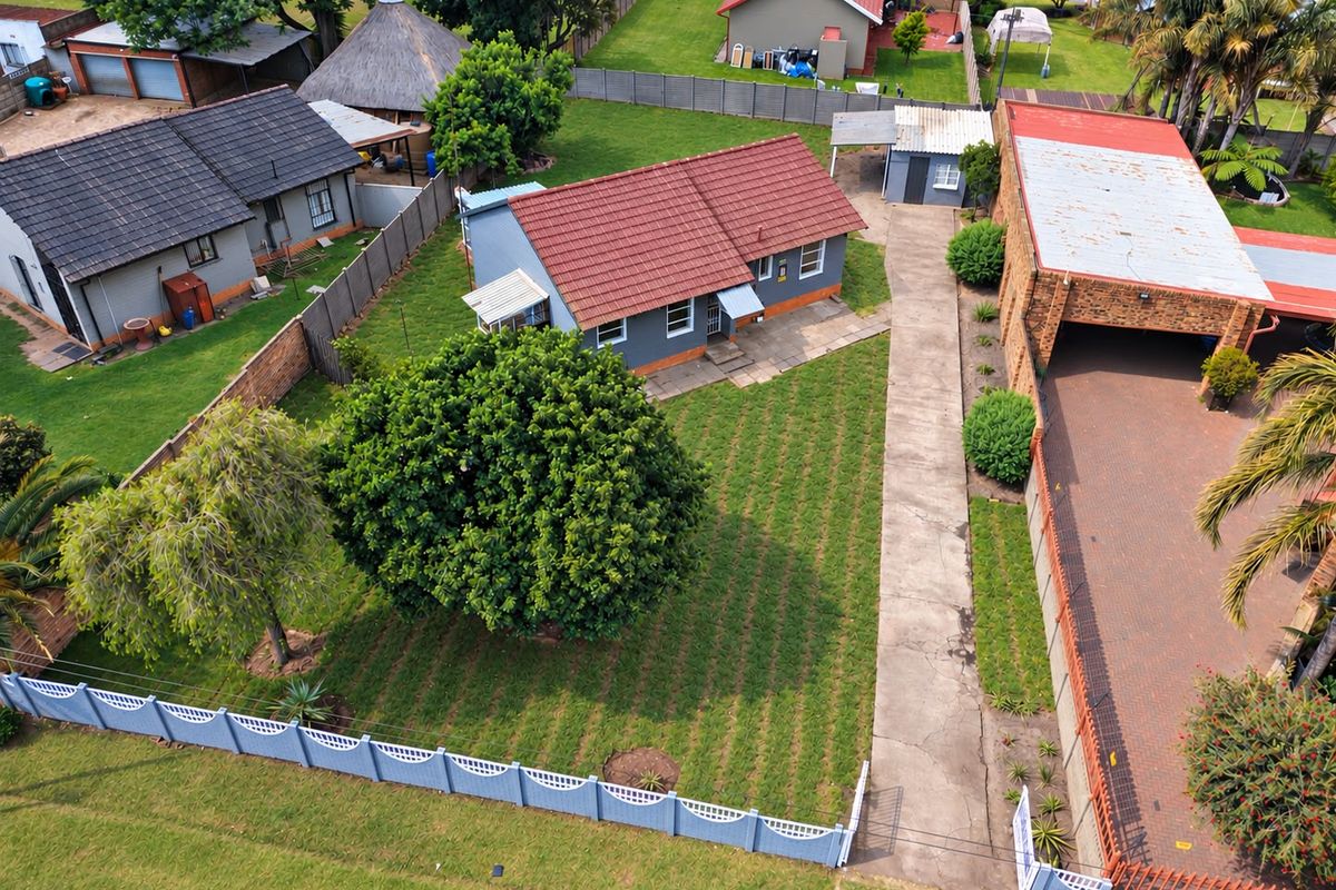 Aerial view of the property showing the main dwelling, large garden, concrete driveway, and detached outbuilding within a fenced residential plot.