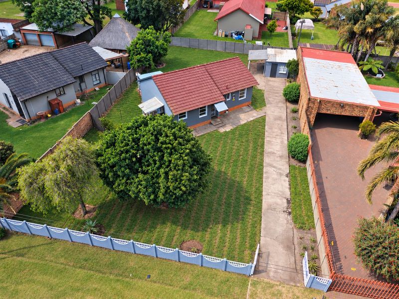 Aerial view of the property showing the main dwelling, large garden, concrete driveway, and detached outbuilding within a fenced residential plot.