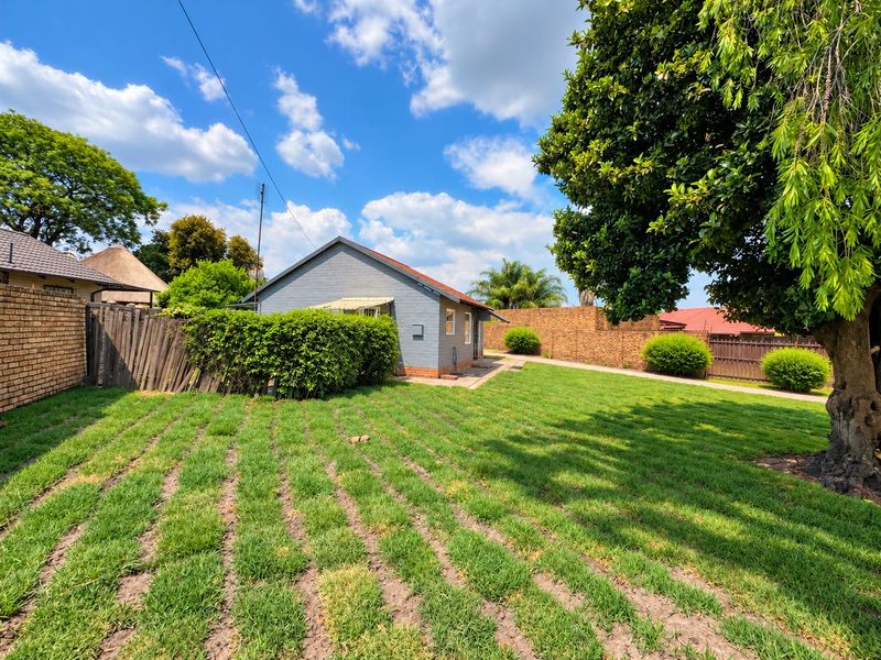 A spacious rear garden featuring a well-maintained lawn, mature trees, and a painted brick exterior facade with a tiled roof.