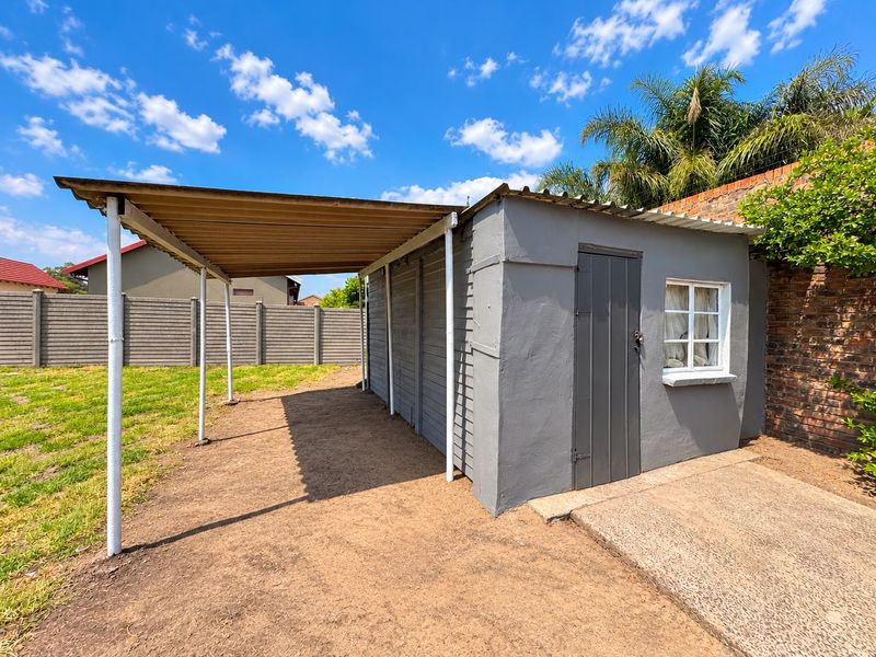 A detached utility structure featuring a covered carport area, concrete apron, and corrugated roofing, situated within a fenced garden setting.