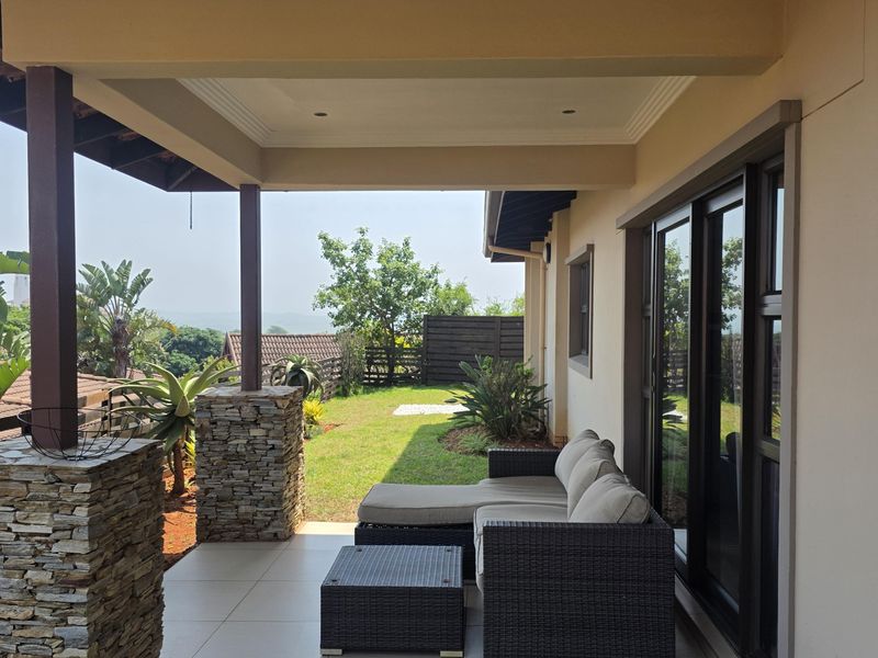 A covered patio featuring stone-clad pillars, tiled flooring, and exposed timber rafters, providing seamless access to the garden through large sliding glass doors.