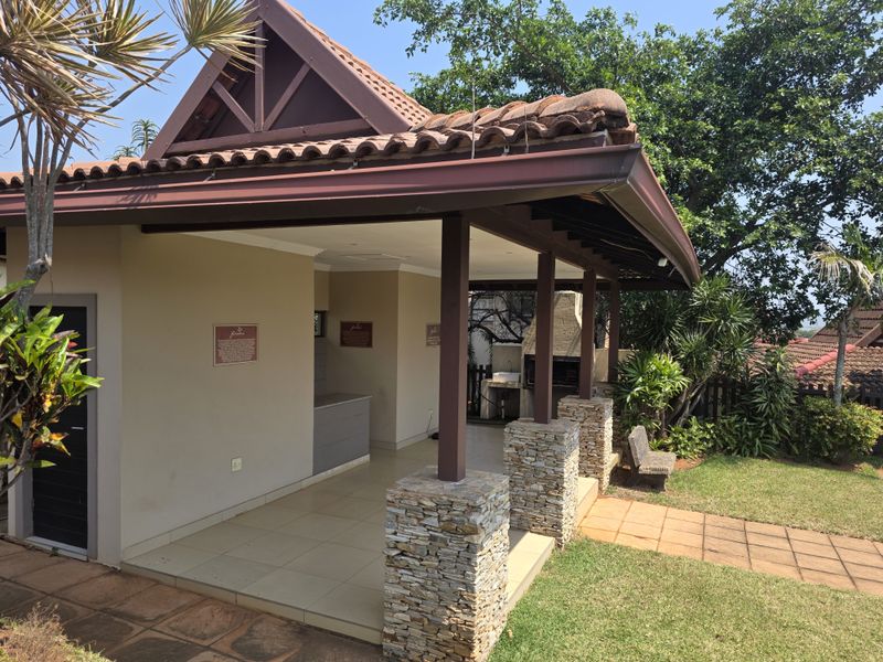 A covered outdoor patio area featuring stone-clad pillars, a built-in braai, and an integrated sink, overlooking a landscaped garden.