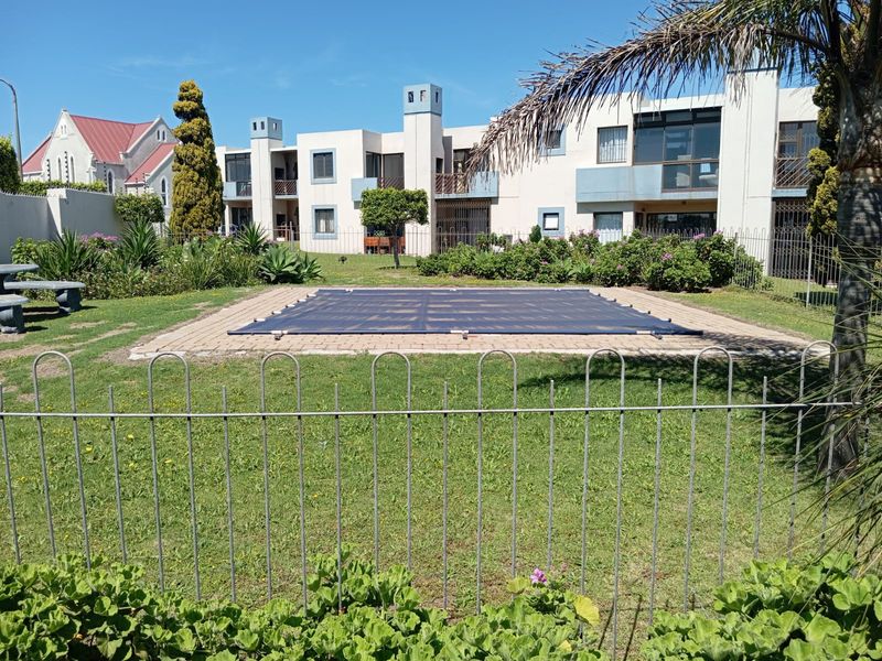 Communal outdoor area featuring a swimming pool with a cover, surrounded by a paved deck, landscaped garden, and secure metal perimeter fencing.