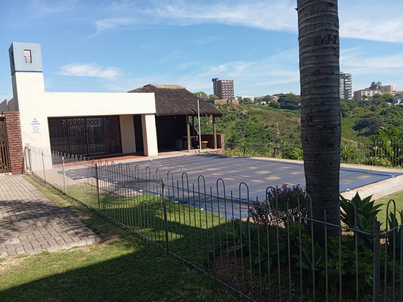 Outdoor pool area featuring a swimming pool with a cover, a thatched lapa, and metal security fencing, set against a scenic hillside backdrop.