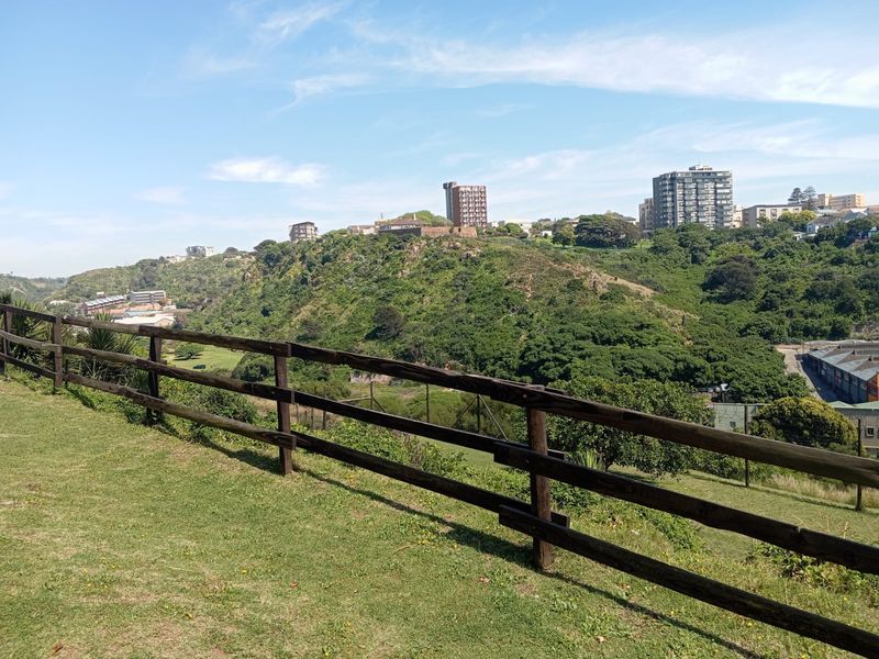 An elevated outdoor view overlooking a lush green valley and surrounding urban landscape, framed by a timber post and rail fence.