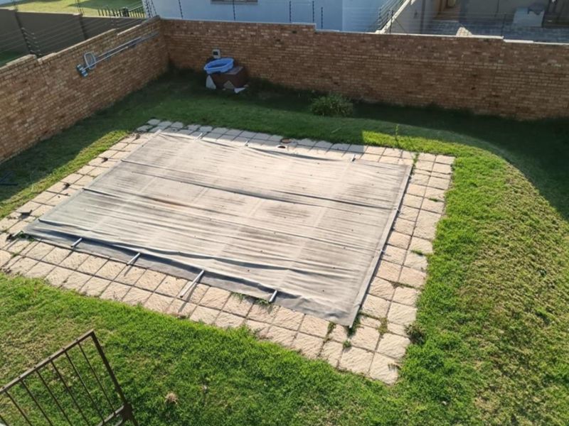 An elevated view of a backyard swimming pool featuring a pool cover, paved surround, and enclosed by a brick boundary wall with electric fencing.
