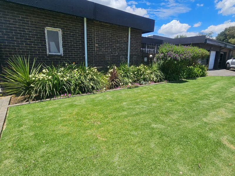 A well-maintained front exterior view featuring a brick facade, a neat lawn, and established garden beds along the driveway.