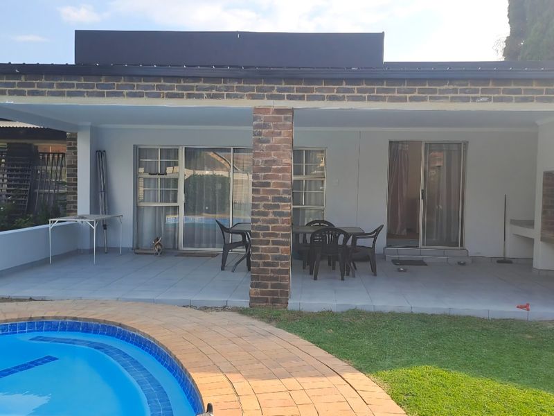 Covered patio area featuring brick pillars, tiled flooring, and a built-in braai, overlooking a swimming pool and garden.