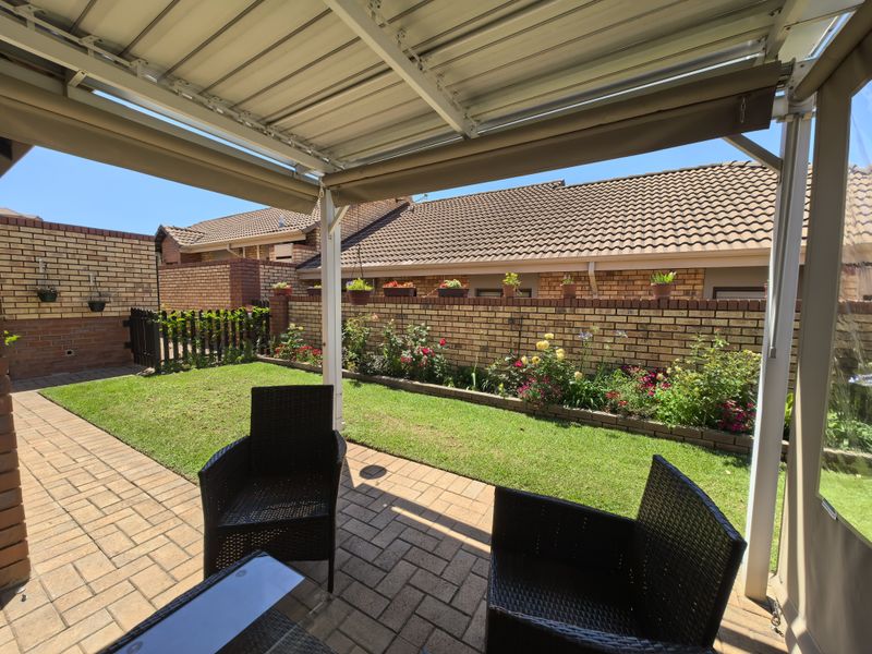 A well-maintained covered patio featuring paved flooring, retractable blinds, and a neat garden with a lawn and brick boundary wall.