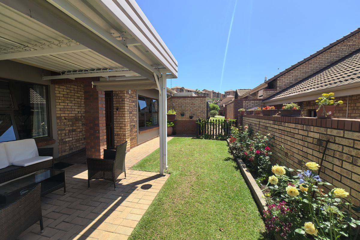 A well-maintained rear exterior featuring a covered patio with paved flooring, a neat lawn, and established garden beds along a brick boundary wall.