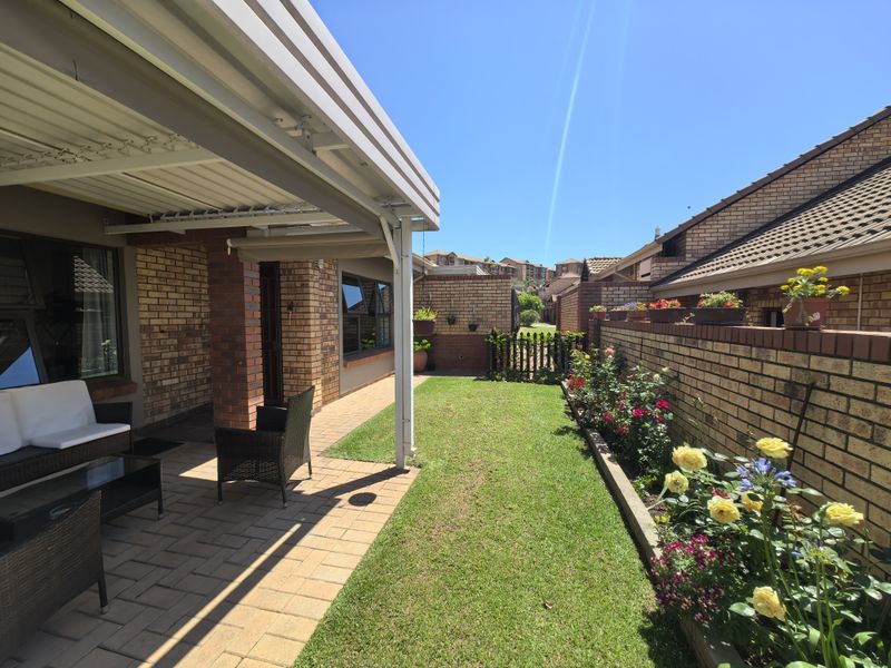 A well-maintained rear exterior featuring a covered patio with paved flooring, a neat lawn, and established garden beds along a brick boundary wall.